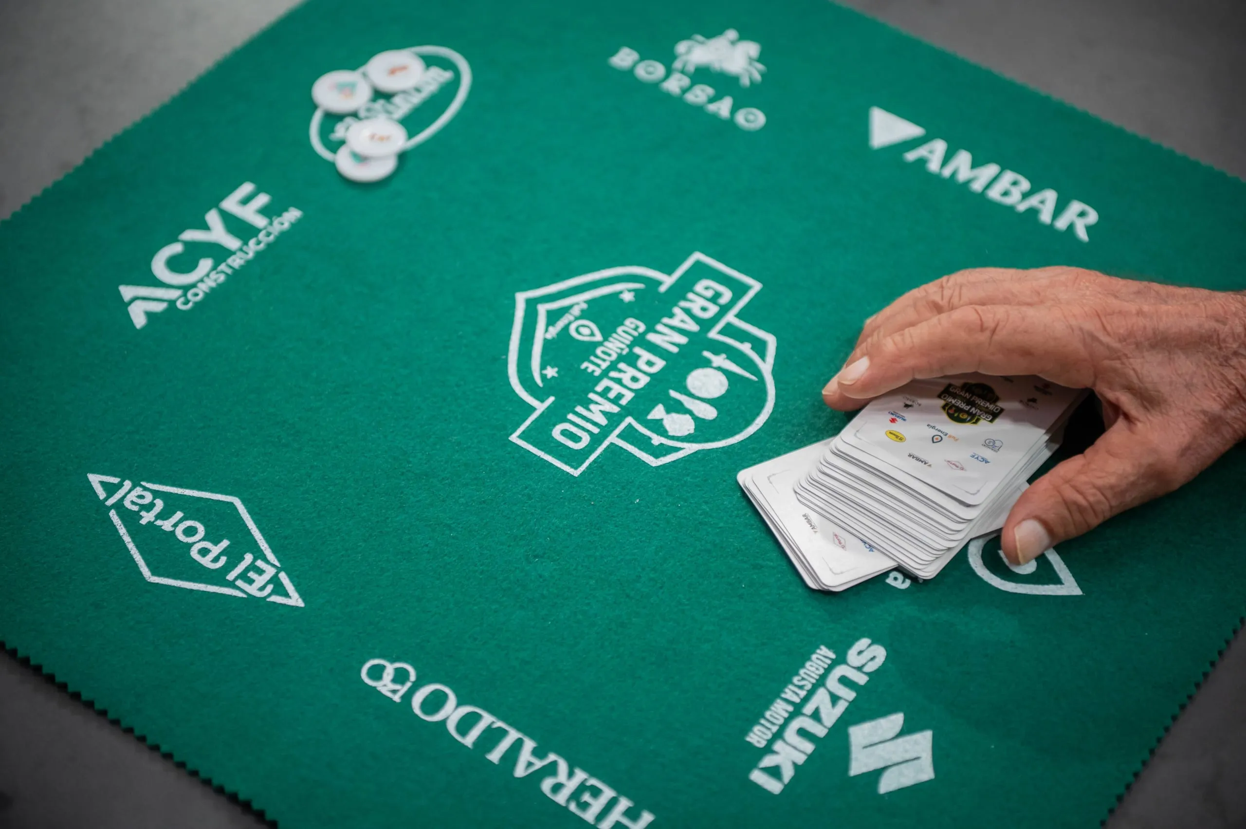 Una mano recoge la baraja sobre el tapete verde del Gran Premio, con el escudo del torneo impreso en el centro.